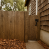Wood fence running perpendicular into the shingled wall of a hillside home with leaf litter piled at the base