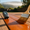Brush applying penetrating stain to a redwood deck board with a moisture meter resting nearby in morning sun